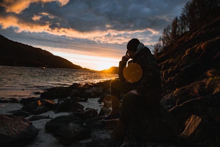 Eine Person in dunkler Kleidung sitzt auf Felsen am Meer und blickt in einen dramatischen Sonnenuntergang – aufgenommen von Julian Haberl | JH-Creative.