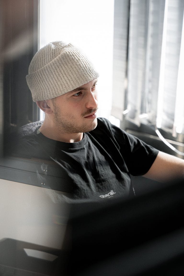 Mann mit Wollmütze sitzt nachdenklich am Fenster, modernes Porträt im Gegenlicht – fotografiert von Julian Haberl