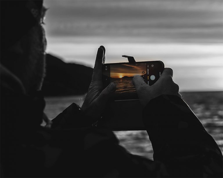 Person fotografiert Sonnenuntergang mit dem Smartphone – Farbakzent im Display vor monochromer Kulisse