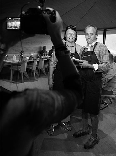 Zwei Personen in Schürzen posieren in einem Alm-Restaurant in Tirol für ein Foto, während ein Fotograf sie aufnimmt.