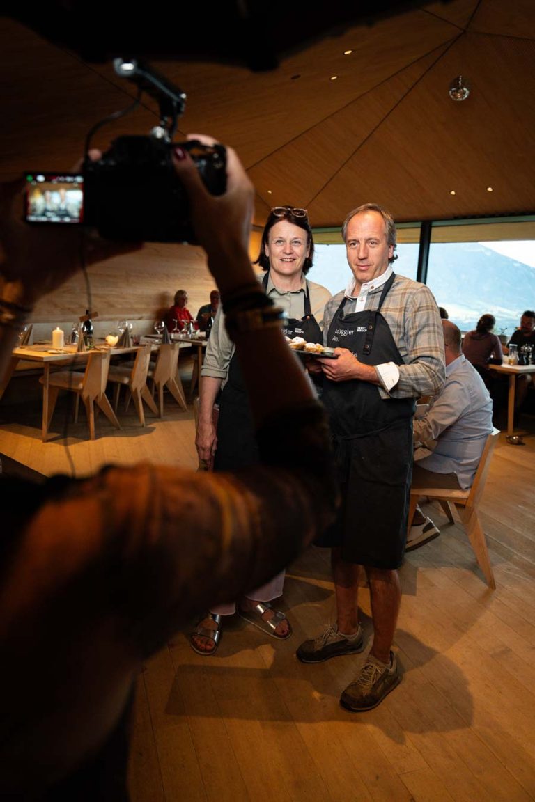 Zwei Personen in Schürzen posieren in einem Alm-Restaurant in Tirol für ein Foto, während ein Fotograf sie aufnimmt.